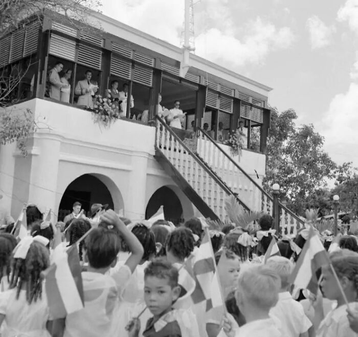 Children waving flags at a public event with dignitaries on a balcony, capturing daily life across the globe in the 50s.