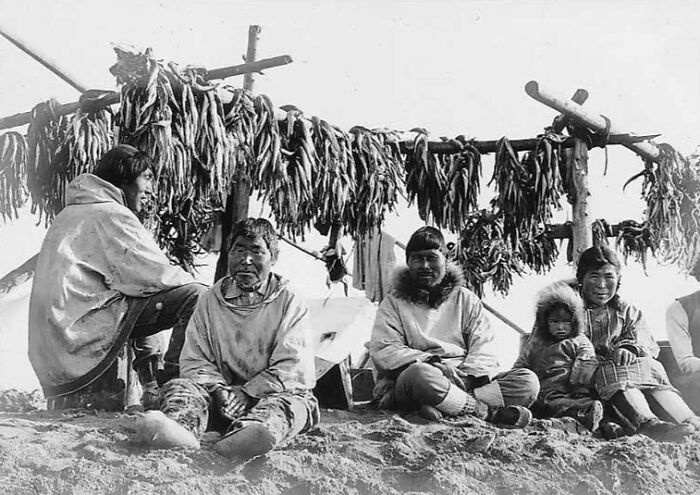 Historic photo of Inuit family sitting by drying fish racks, showcasing Inuit spirit and traditional lifestyle in a snowy environment.
