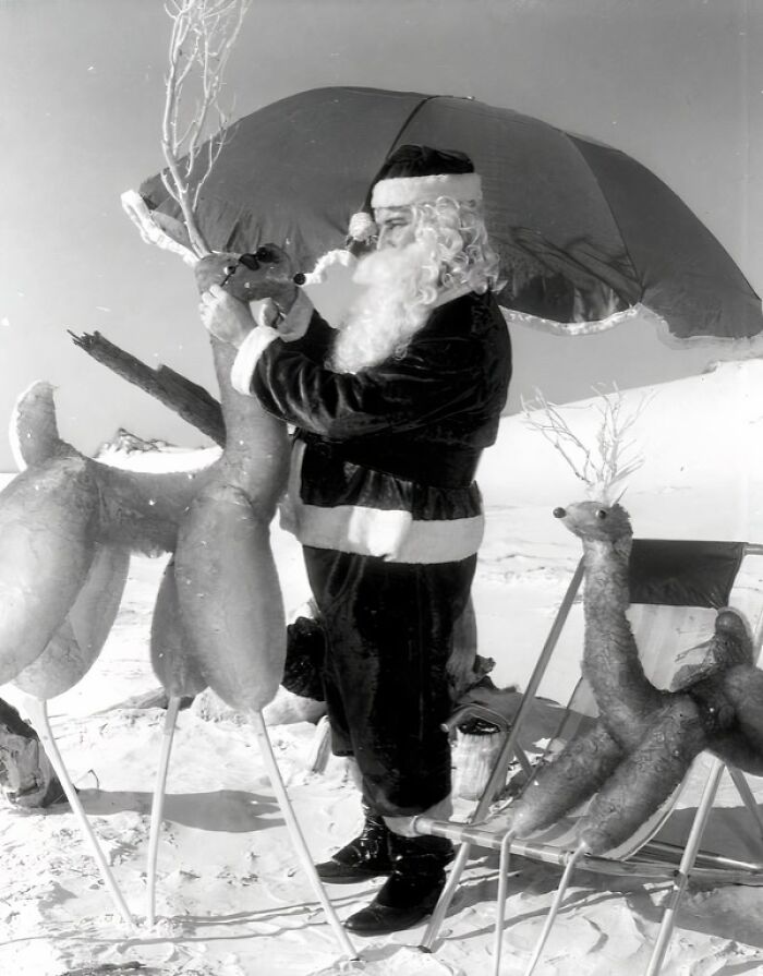 Santa Claus on a Florida beach adjusting reindeer decorations under an umbrella in a 1950s everyday life scene.