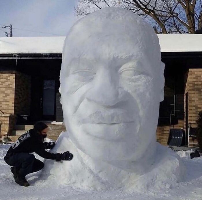 Person sculpting a giant, detailed snow head sculpture outside a house, showing unhinged cursed images creativity.