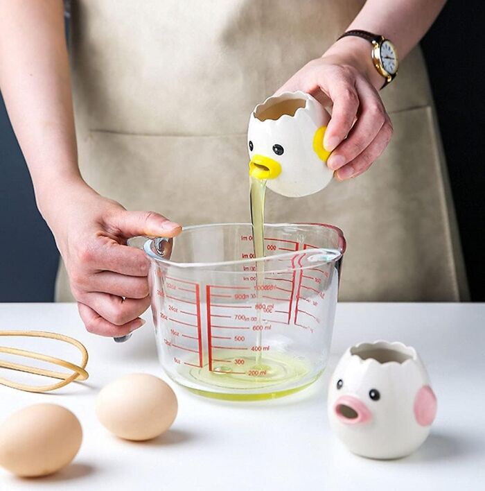 Person cracking egg using a weird kitchen invention shaped like a chick to pour egg white into a measuring cup.