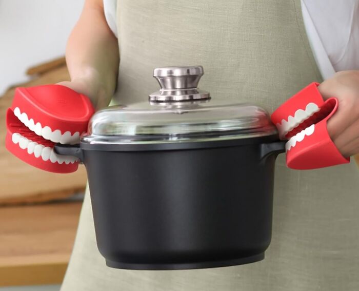 Person wearing quirky red chattering teeth oven mitts holding a black pot with a glass lid in the kitchen.