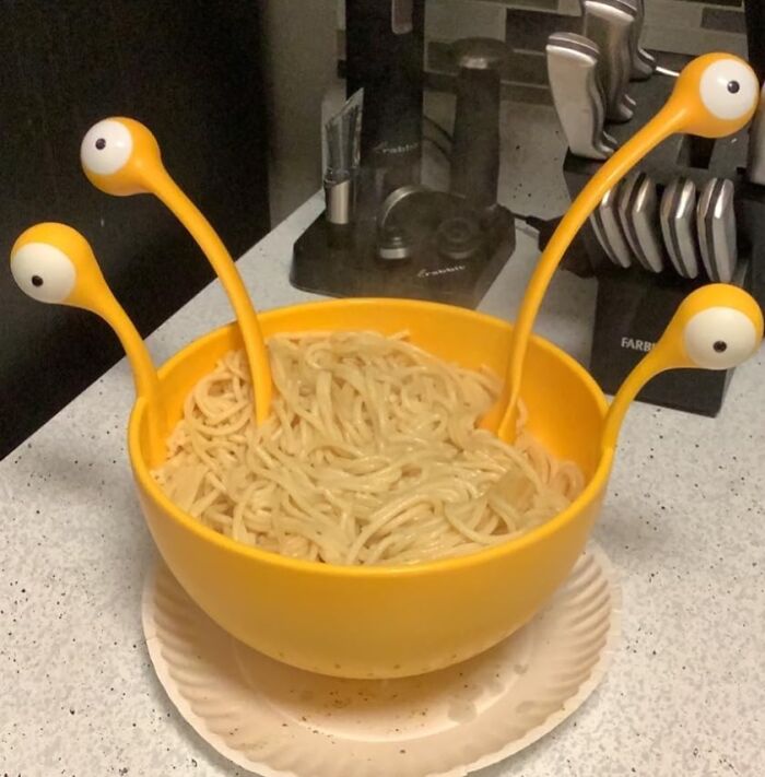 Yellow bowl with cooked spaghetti and quirky weird kitchen inventions shaped like eyes on flexible stalks.