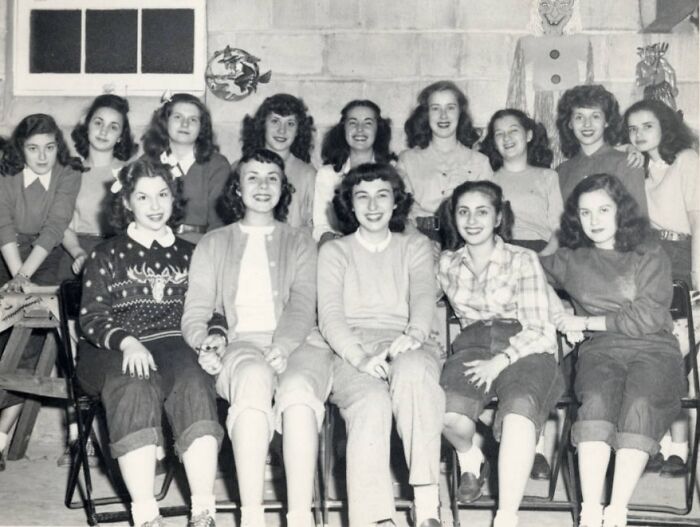 Group of teenage girls posing together inside a school classroom, historical black and white photo from World War II era.