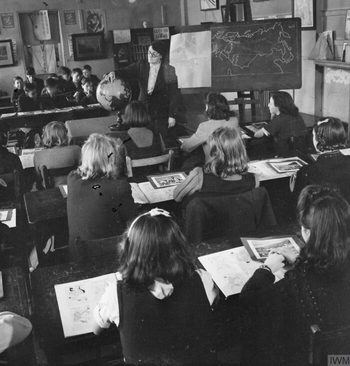 World War II school classroom with teacher pointing at globe and students studying maps and geography lessons.