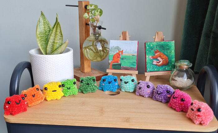 Colorful crochet frogs lined up on a wooden table with small plant and cat paintings in the background.