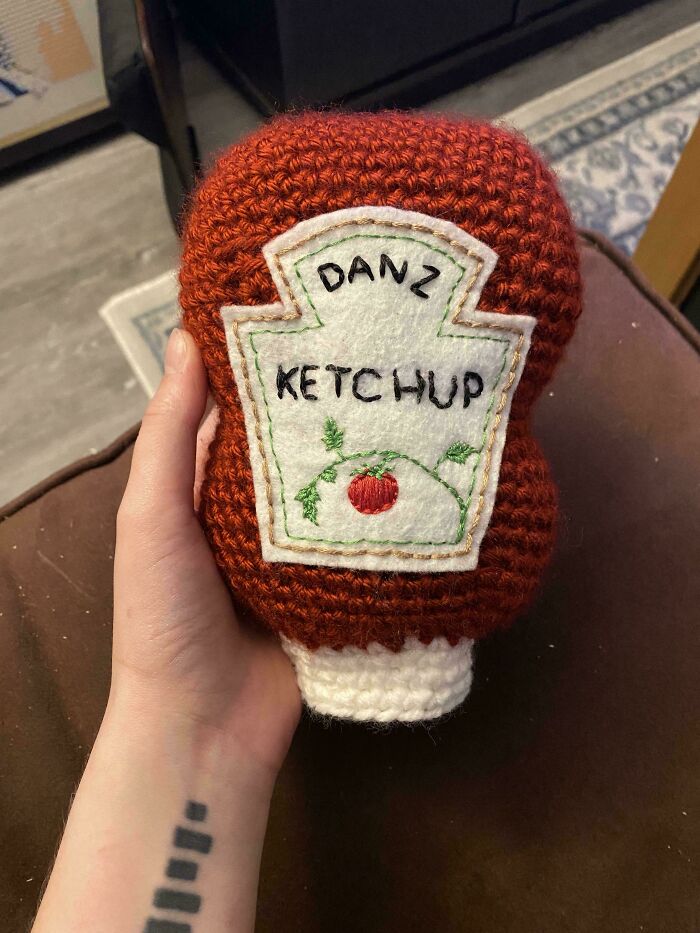 Hand holding an intricately crocheted ketchup bottle with embroidered label, showcasing crochet enthusiasts’ creative work.