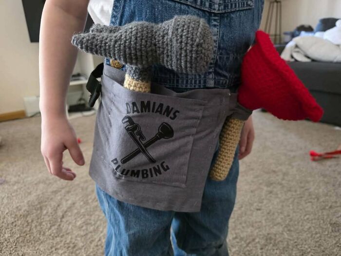 Crochet enthusiast holding handmade crochet tools including a gray hammer and red plunger in a tool belt.