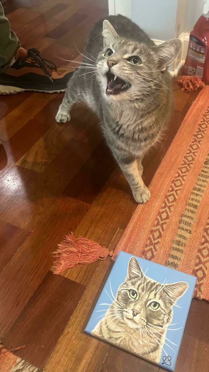 Tabby cat meowing loudly indoors near a painted tile of a similar cat on a wooden floor with a rug nearby.