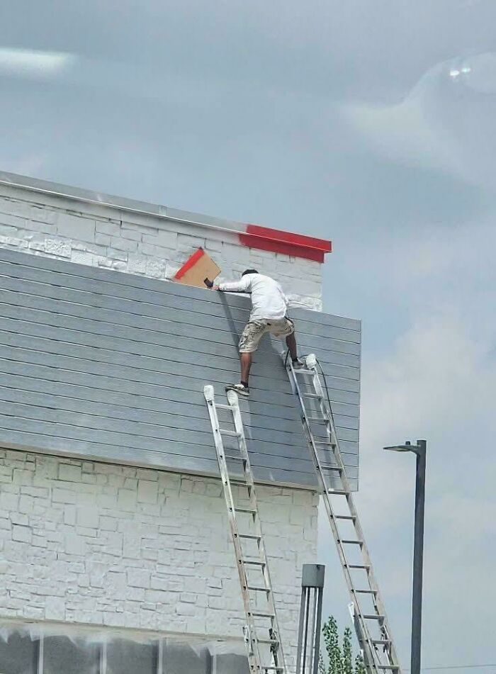 Employee blatantly ignoring safety protocol by standing unsafely on two ladders to work on building exterior.