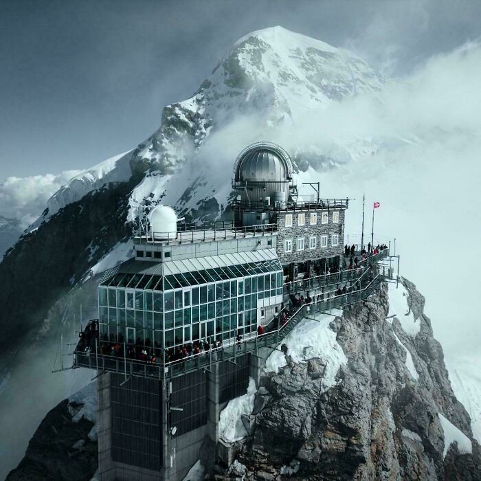 Modern architectural feat perched on a snowy mountain peak with glass and stone structures against a cloudy sky.