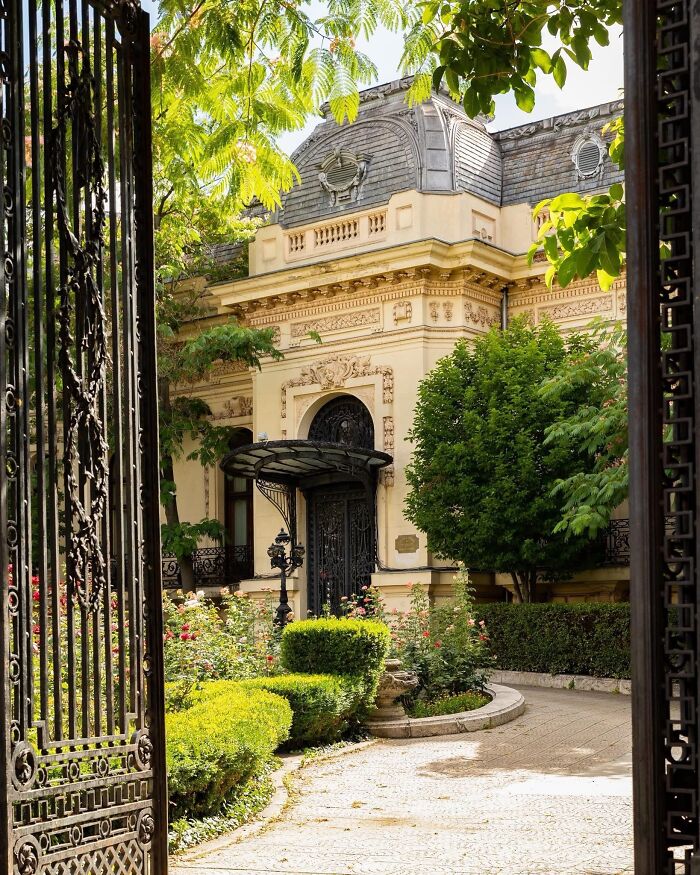 Elegant historic building with detailed architectural feats surrounded by lush greenery and a wrought iron gate entrance.