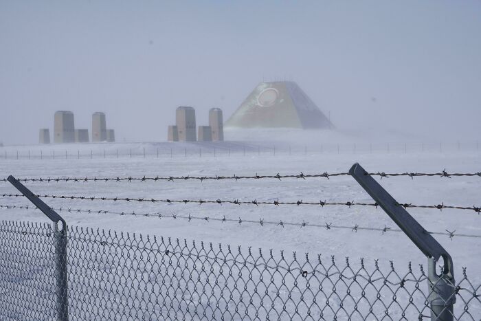 Snow-covered scene with a barbed wire fence and an eerie, pyramid-shaped evil building in the distance.
