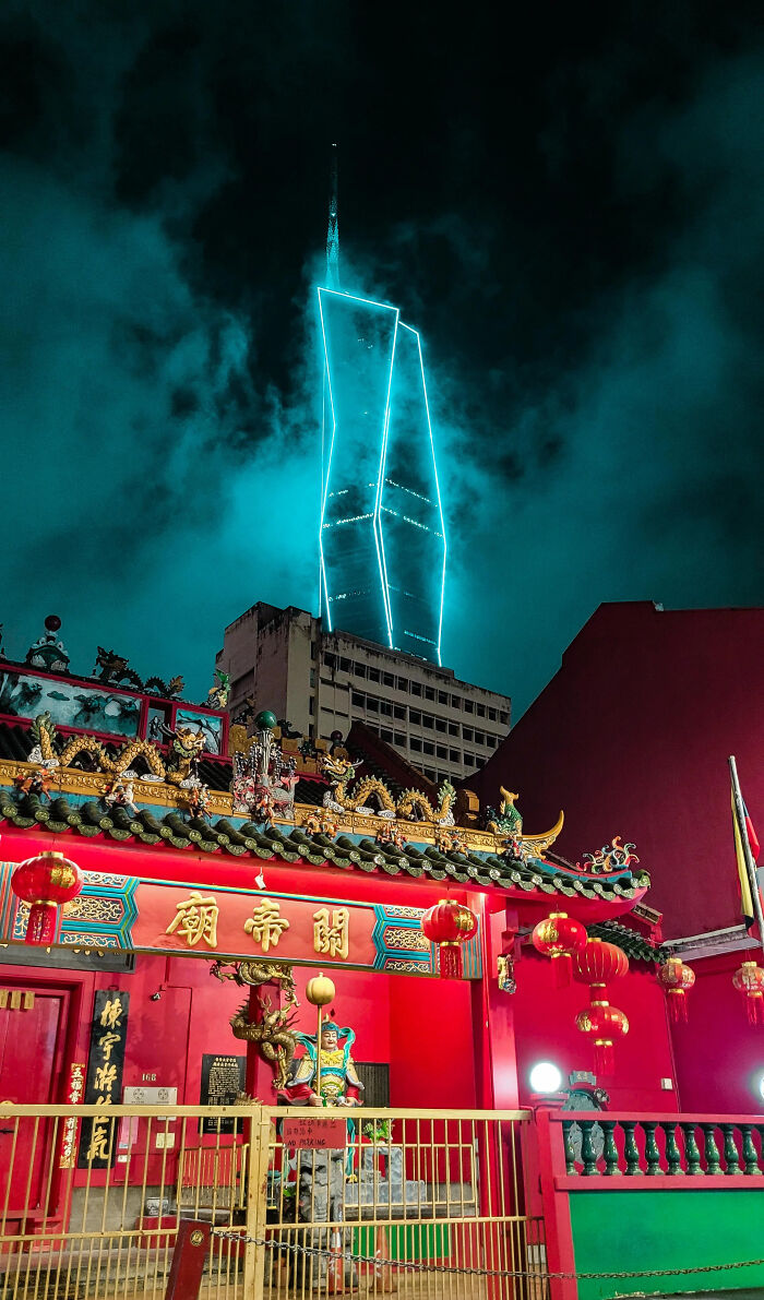 Modern building with glowing outlines looms behind a traditional temple, creating an eerie scene of evil buildings at night.