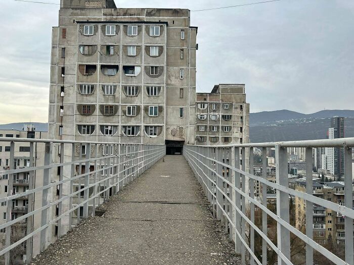 Concrete evil building with ominous dark entrance and narrow elevated walkway in an urban setting with distant hills.