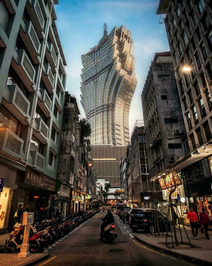 Futuristic evil building towering over narrow city street lined with old apartments and parked scooters at dusk.