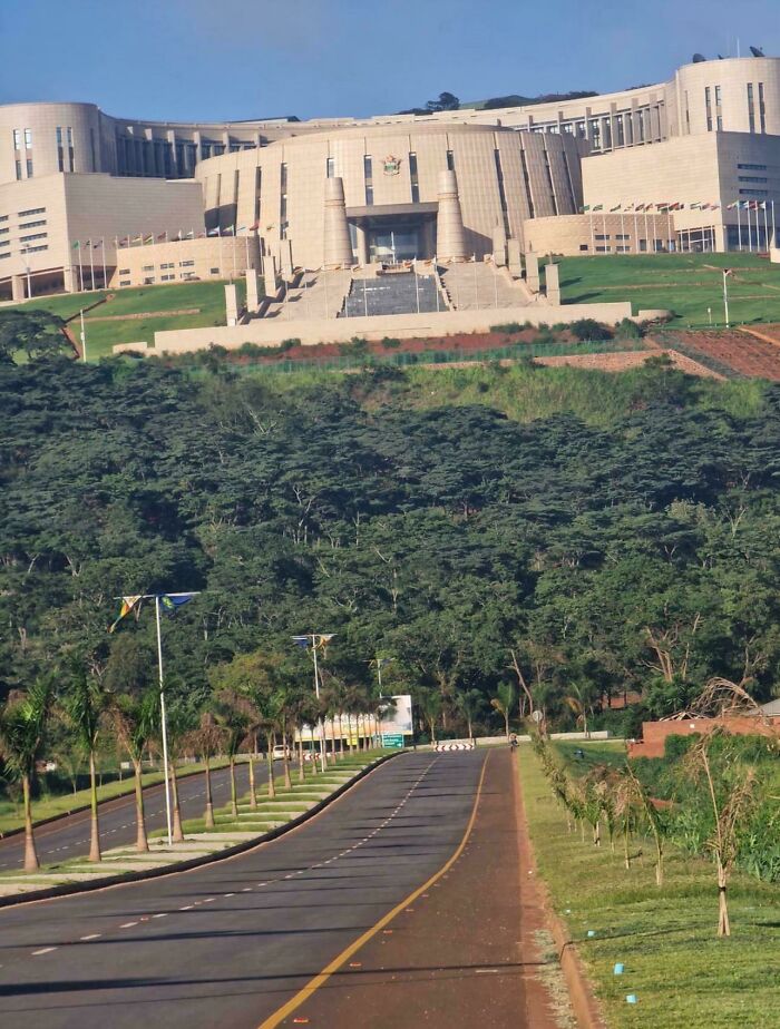 Massive evil building on a hill with a wide stairway entrance surrounded by trees and a long empty road leading up to it.