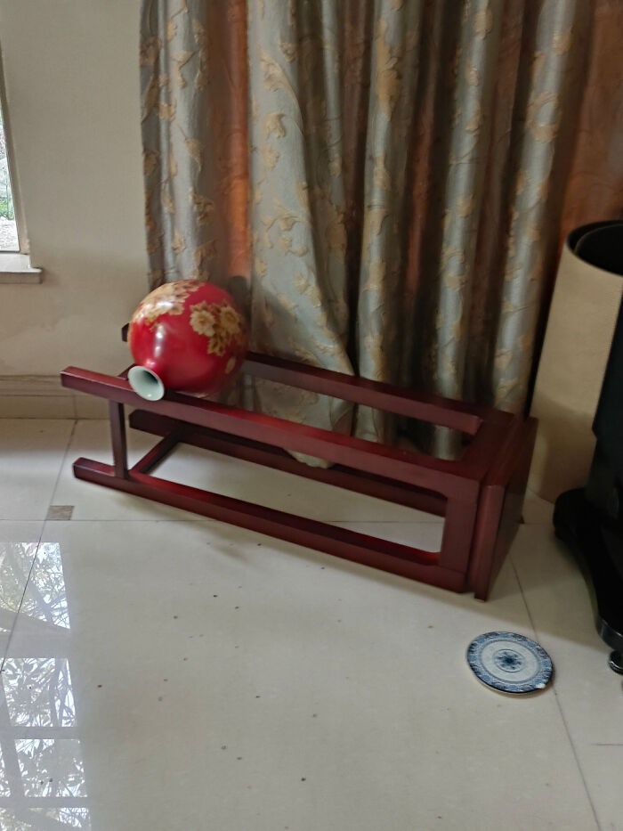 Floral vase lying inside upside-down wooden stand next to a broken plate on floor, a coincidence captured in a photo.
