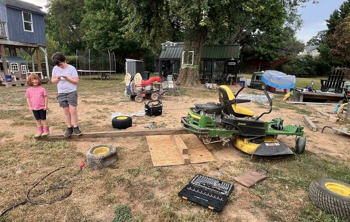 Two kids standing next to a homemade redneck engineer contraption made from a lawnmower and wooden planks outdoors.