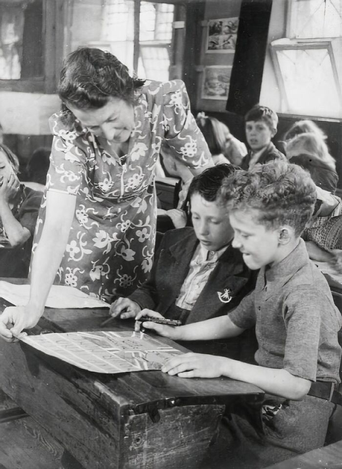Teacher assisting two boys at a wooden desk in a classroom, a historic scene of schools worldwide during World War II.
