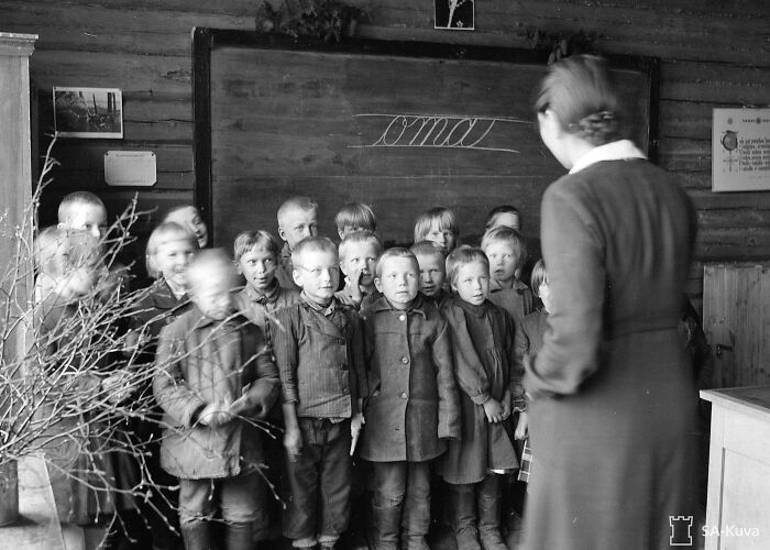 Group of schoolchildren standing inside a classroom during World War II, with a teacher near the blackboard.