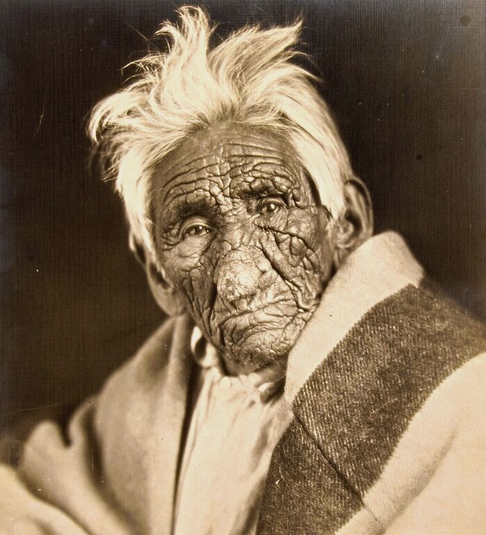 Somber historic photo of an elderly person with deeply wrinkled skin and white hair wrapped in a blanket.