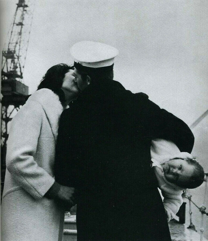 Sailor kissing woman goodbye while holding a child on a ship deck in a somber photo from history.