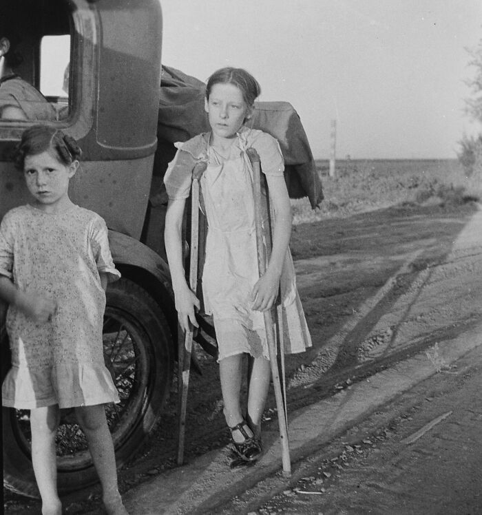 Two young girls stand beside an old car on a dirt road in a somber photo from history showing hardship.