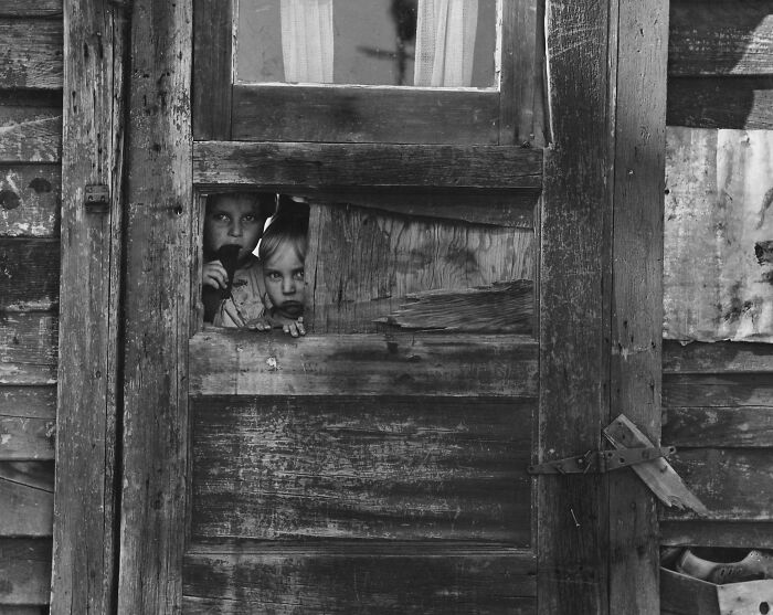 Two children peering through a broken wooden door, a somber photo conveying a gut-wrenching story from history.