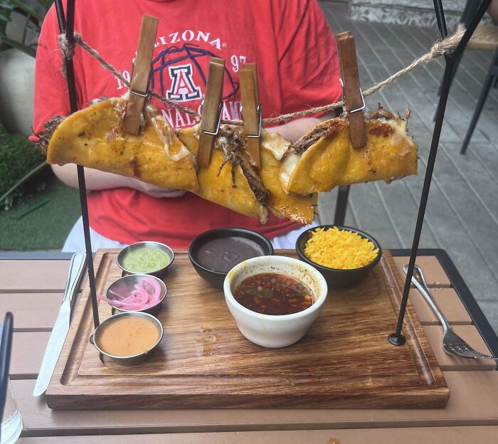 Tacos served hanging on a clothesline with clothespins above a wooden board with sauces and sides in a restaurant.