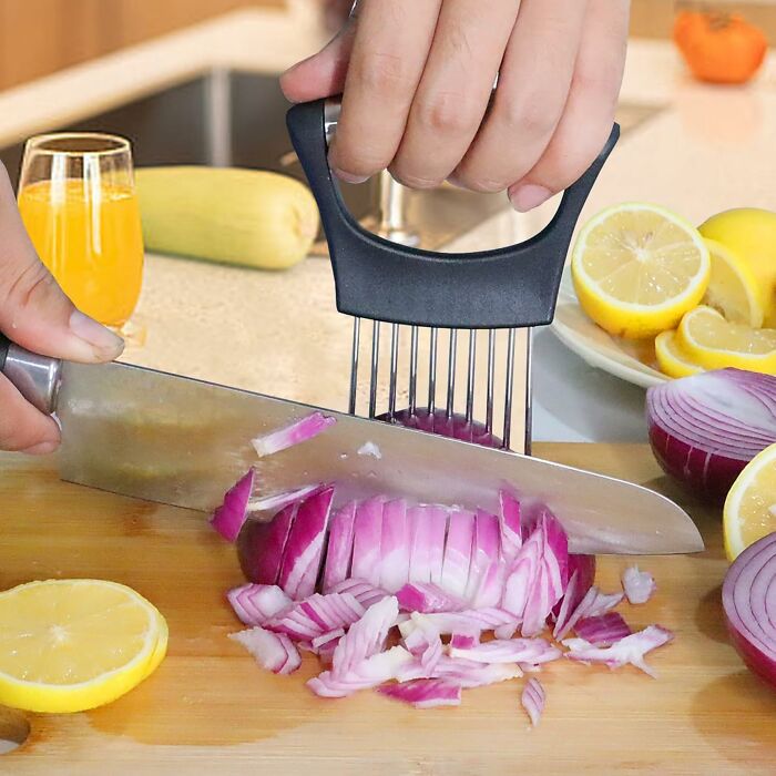 Hand using a weird kitchen invention to hold an onion for even slicing on a wooden cutting board with lemons nearby.