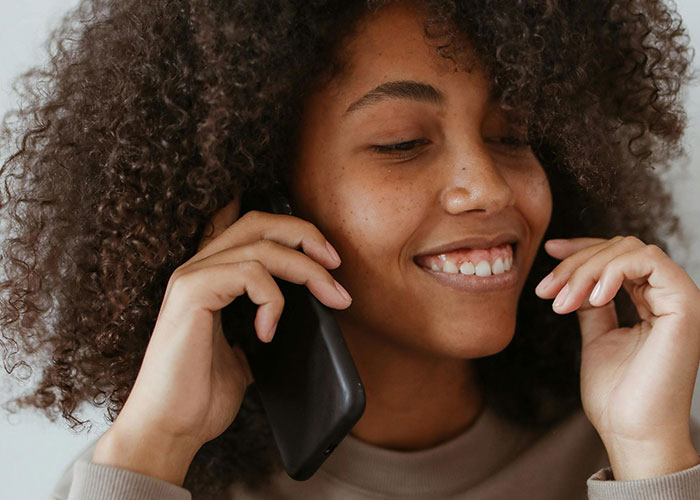 Young woman with curly hair smiling while talking on a phone, illustrating bizarre family habits shared by people.
