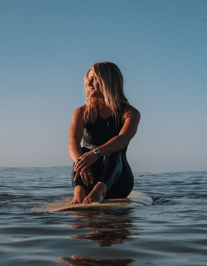 Surf instructor sitting on a surfboard in the ocean during sunset, wearing a black wetsuit and looking to the side. Surf instructor sitting on a surfboard in the ocean during sunset, wearing a black wetsuit and looking to the side.