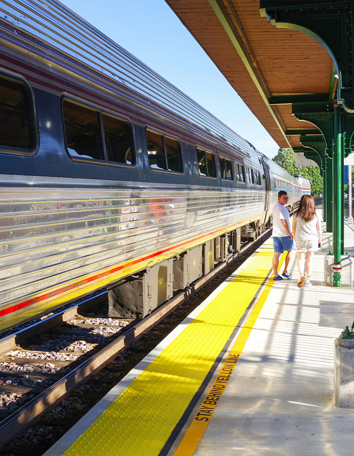 Train at a sunny station platform with two people walking near the yellow safety line, related to surf instructor accident incident. Train at a sunny station platform with two people walking near the yellow safety line, related to surf instructor accident incident.