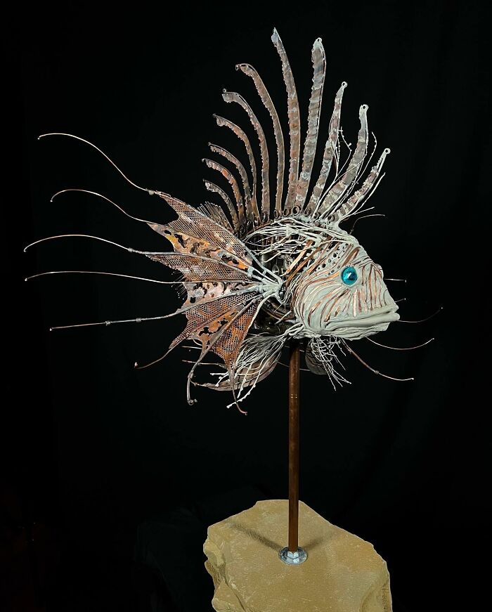 Intricate scrap metal animal sculpture of a fish with detailed fins and bright blue eyes on a stand against black background.