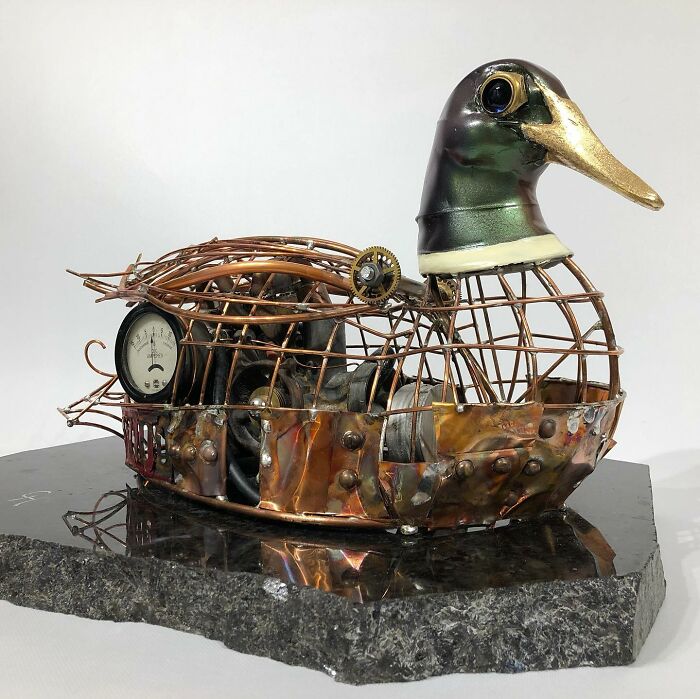 Scrap metal animal sculpture resembling a duck with intricate gears and a gauge, displayed on a stone base.