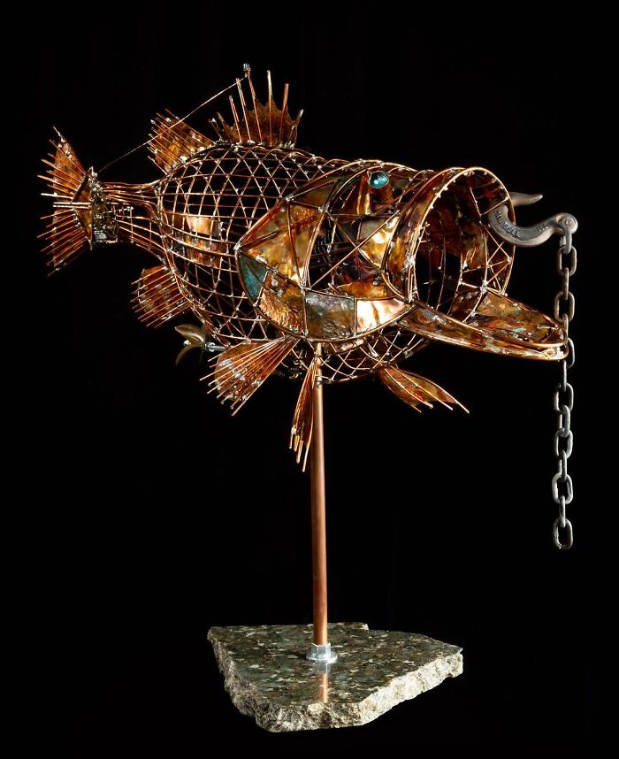 Metal fish sculpture made from scrap materials, showcasing intricate animal sculpture artistry on a stone base against black background.