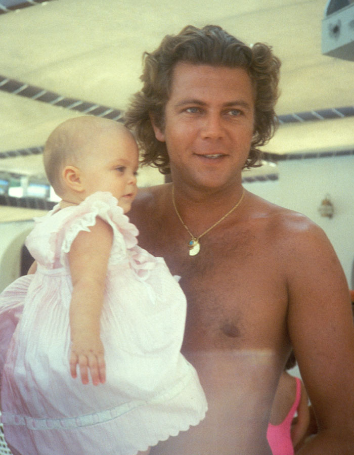 Man holding a baby girl in a pink dress under a shaded outdoor area, related to reclusive billionaire Athina Onassis. Man holding a baby girl in a pink dress under a shaded outdoor area, related to reclusive billionaire Athina Onassis.