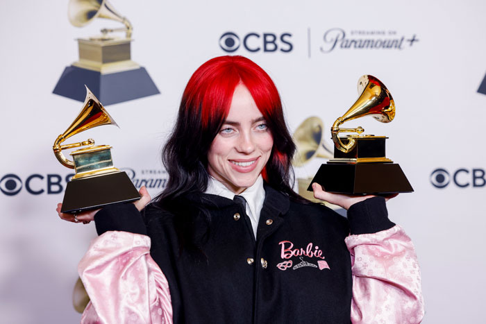 Billie Eilish holding two Grammy awards, smiling with red and black hair at a CBS and Paramount event. Billie Eilish holding two Grammy awards, smiling with red and black hair at a CBS and Paramount event.