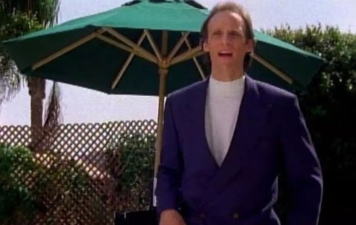 Man in a dark blazer standing outdoors under a green umbrella, with greenery and lattice fence in the background. Man in a dark blazer standing outdoors under a green umbrella, with greenery and lattice fence in the background.