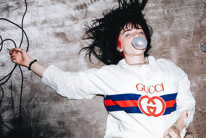 Billie Eilish lying down wearing a Gucci sweatshirt and blowing a bubble with gum, sparking nepo baby fan discussions. Billie Eilish lying down wearing a Gucci sweatshirt and blowing a bubble with gum, sparking nepo baby fan discussions.