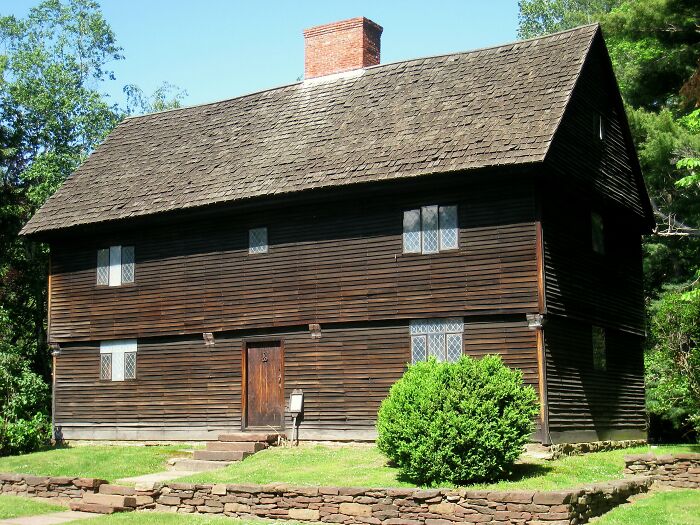 Historic wooden home with a steep roof and chimney, surrounded by greenery, showcasing America's architectural heritage.