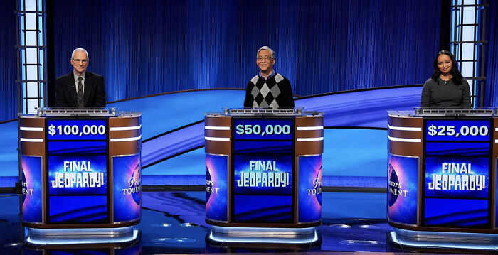 Three Jeopardy contestants standing behind podiums with Final Jeopardy screens showing their scores on the quiz show set. Three Jeopardy contestants standing behind podiums with Final Jeopardy screens showing their scores on the quiz show set.