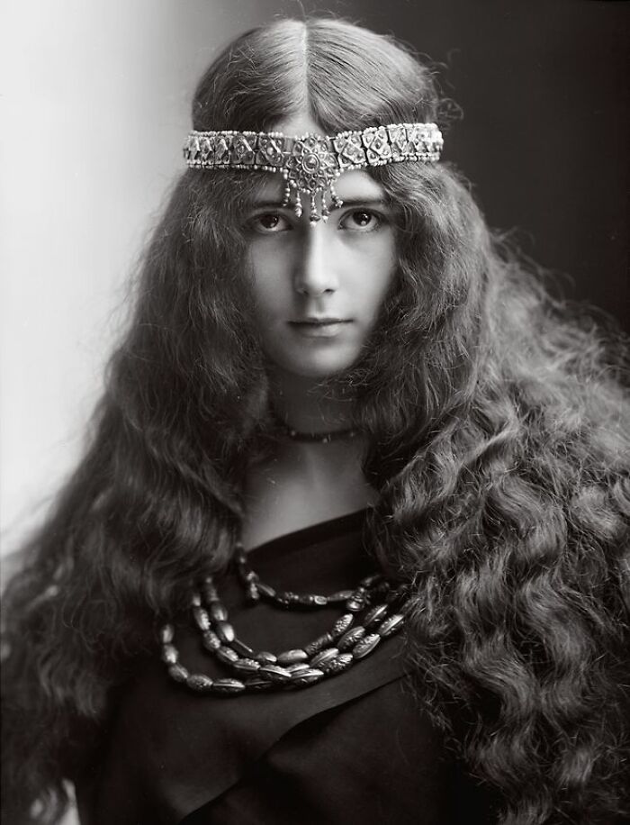 Century-old photo of a woman with long wavy hair and ornate headpiece embodying timeless beauty and elegance.