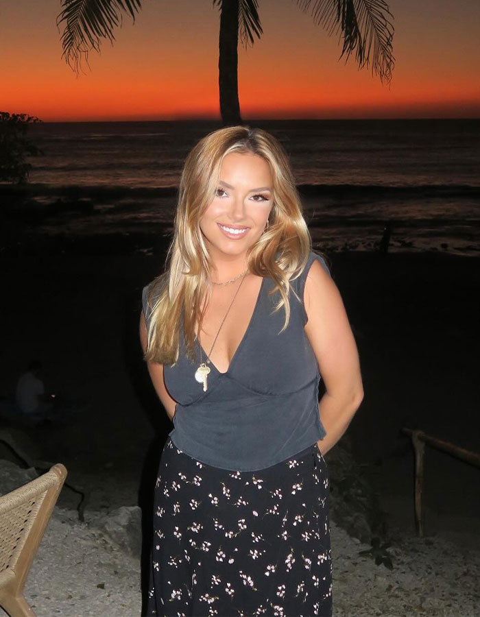 Young woman smiling outdoors at sunset with palm trees, related to Coldplay fan who exposed cheating CEO by filming kiss cam. Young woman smiling outdoors at sunset with palm trees, related to Coldplay fan who exposed cheating CEO by filming kiss cam.