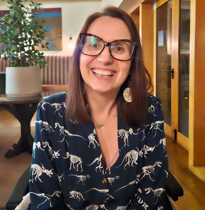 Smiling woman wearing glasses and dinosaur skeleton print shirt, sitting indoors in a cozy room. Smiling woman wearing glasses and dinosaur skeleton print shirt, sitting indoors in a cozy room.