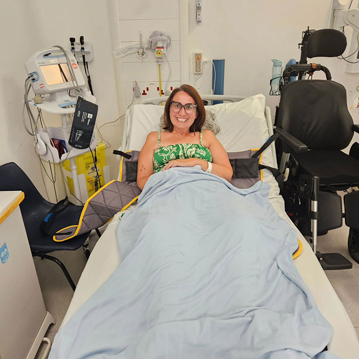 Woman lying in hospital bed, smiling, surrounded by medical equipment in a clinical room setting. Woman lying in hospital bed, smiling, surrounded by medical equipment in a clinical room setting.