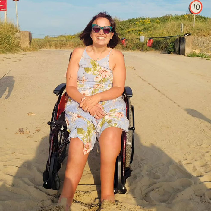 Woman sitting in a wheelchair on a sandy path, wearing sunglasses and a floral dress, smiling in sunlight. Woman sitting in a wheelchair on a sandy path, wearing sunglasses and a floral dress, smiling in sunlight.
