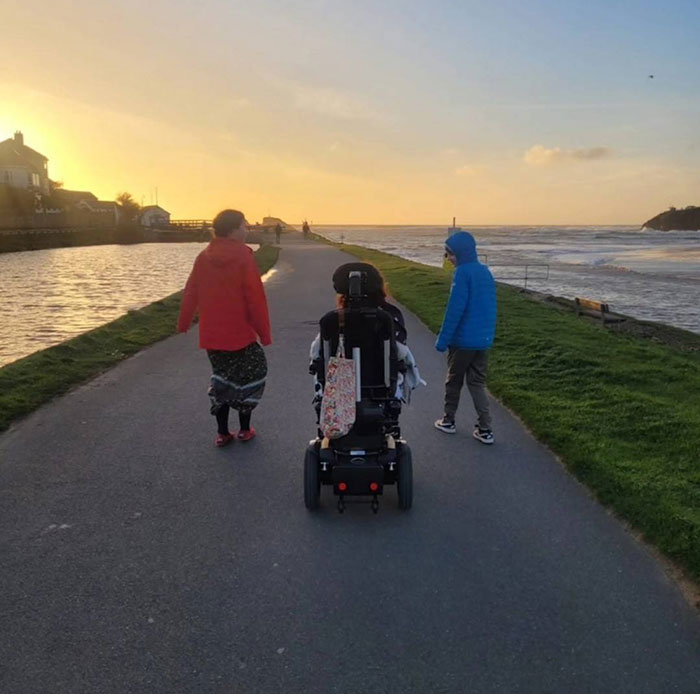 Three people including a woman in a wheelchair walking near the sea at sunset, illustrating a mom protecting kids from struggle. Three people including a woman in a wheelchair walking near the sea at sunset, illustrating a mom protecting kids from struggle.