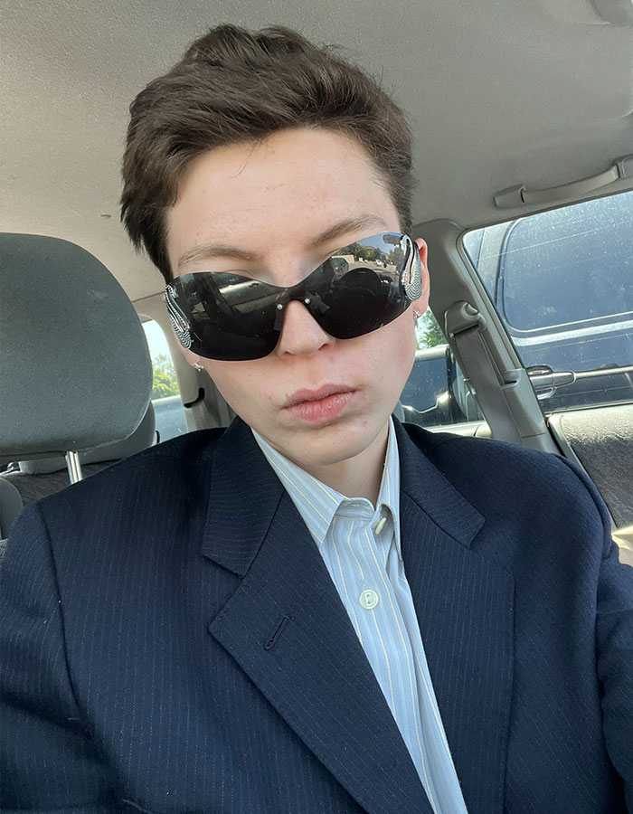 Person with short hair wearing dark sunglasses and a suit jacket sitting inside a car, related to model detransition story. Person with short hair wearing dark sunglasses and a suit jacket sitting inside a car, related to model detransition story.